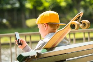 Telemedien: Ein Junge sitzt draußen auf einer Parkbank, ein Skateboard neben sich aufgestellt, und beschäftigt sich mit einem Smartphone.